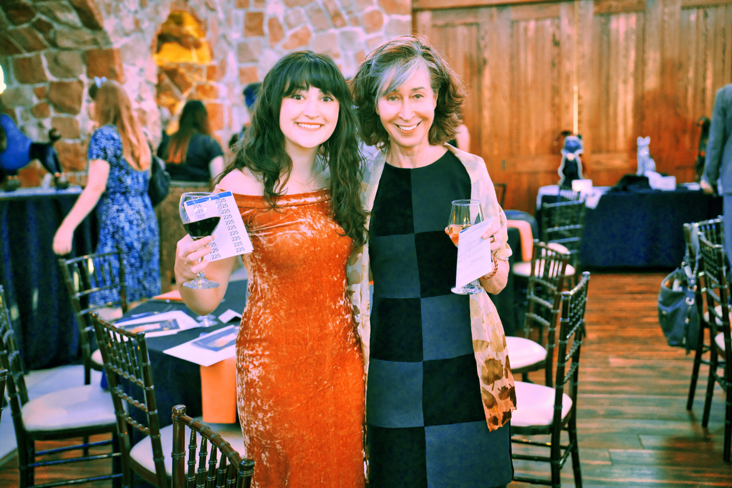 Guests at TOPC 2023. Woman on left is wearing a red dress. Woman on right is wearing a black checkered dress. Both women are holding wine glasses and smiling for a photo.